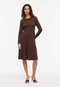 Robe marron à manches longues avec un corsage ajusté, une jupe à volants et un col rond. Fabriquée en matière texturée, associée à des talons ouverts noirs.