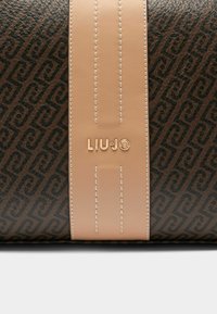 Brūna ādas rokas soma ar atkārtotu ģeometrisku rakstu, gludu bēša krāsas lenti ar šuvumiem un metālisku logo detaļu.