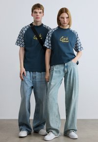 Δύο μοντέλα φορούν όμοια σκούρα μπλε-πράσινα μπλουζάκια με μοτίβα στα μανίκια και κίτρινο λογότυπο. Και τα δύο συνδυάζουν με άνετα ανοιχτό μπλε τζιν και λευκά αθλητικά παπούτσια.