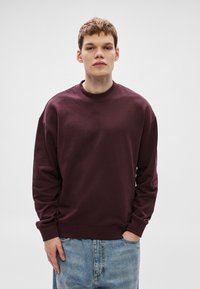 Bordó pulóver kerek nyakkivágással, bordázott mandzsettákkal és derékrésszel. Finom logózás a mellkason, laza fazon. Farmerrel viselve.