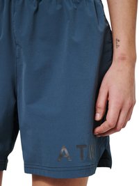 Close-up van een persoon die een blauwe korte broek draagt met "ATW" gedrukt vlak bij de zoom en die zijn rechterhand toont met een kleine polstattoo.