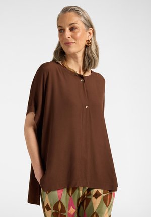 Femme aux cheveux gris mi-longs, portant un haut marron ample et un pantalon à motifs géométriques colorés, regardant sur le côté, les mains dans les poches.