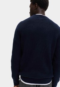 Suéter texturizado azul marino con cuello redondo y dobladillo acanalado, que presenta mangas largas y material suave, llevado sobre una camisa blanca.