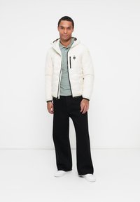 Doudoune de couleur claire avec capuche, design matelassé et poches zippées, assortie à un polo vert et à un pantalon noir. Baskets blanches.