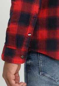 Chemise à carreaux rouge et marine avec des poignets à boutons et un détail de logo, fabriquée à partir d'un tissu texturé, associée à un jean bleu clair.
