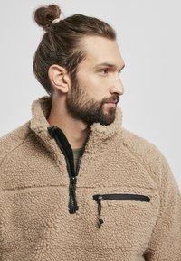 Beige fleece pullover med høj krave, halv lynlås fortil og en lynlåslomme på brystet med sort hardware. Blødt, struktureret stof.