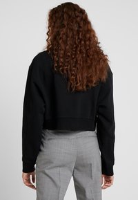Sudadera negra corta con mangas largas, tejido texturizado y un diseño simple. Llevada sobre pantalones de cuadros grises.