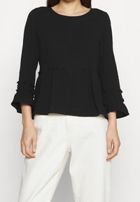 Schwarzes, strukturiertes Peplum-Top mit langen Ärmeln und Rüschendetails. Der runde Halsausschnitt ergänzt den lockeren Schnitt.