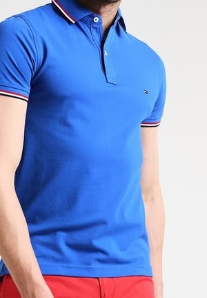 Mand iført en blå poloshirt med rød, hvid og marineblå stribet kant på ærmer og krave, kombineret med røde bukser.