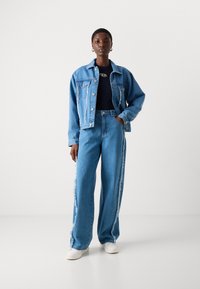 Denimjacke und weite Denimhosen in Hellblau. Die Jacke hat Fransenakzente; die Hosen haben kontrastierende Seitenstreifen und eine lässige Passform.