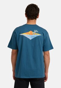 Mann trägt ein blaues T-Shirt mit einem bunten Sonnenuntergang, Ozeanwellen und einem Palmenbaum-Grafik auf dem Rücken, stehend vor einem schlichten weißen Hintergrund.
