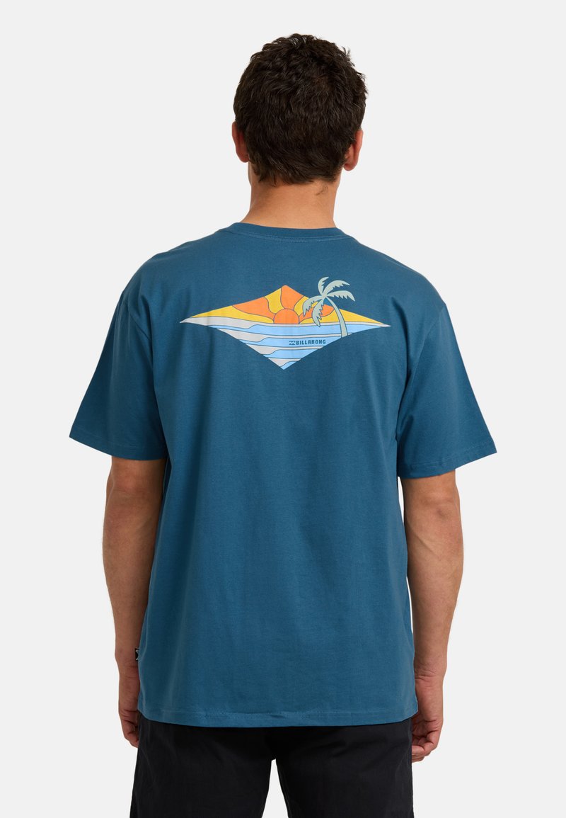 Mann trägt ein blaues T-Shirt mit einem bunten Sonnenuntergang, Ozeanwellen und einem Palmenbaum-Grafik auf dem Rücken, stehend vor einem schlichten weißen Hintergrund.
