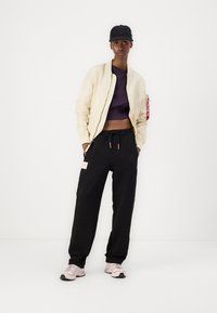 Beige bomberjakke, lilla kort topp, svarte joggebukser med snøring og sidelommer, og lys rosa sko. Enkelt design og afslappet fit.