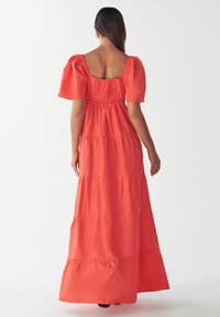 Koralīgas maksi kleita ar pūkainām piedurknēm, kvadrātveida aizmuguri, elastīgu jostu un slāņotu svārku. Gluda auduma ar plūstošu siluetu.