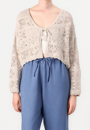Vrouw draagt een beige gehaakte cardigan die aan de voorkant is gestrikt over een witte top en losse blauwe broek met trekkoord, tegen een effen achtergrond.