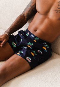 Marinefarbene Schwimmshorts mit einem Bund, die bunte Muster von Campingbussen, Palmen und Inseln zeigen, vor einem glatten Hintergrund.