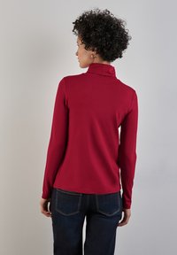 Röd långärmad topp med hög krage, tillverkad av slät tyg, kombinerad med mörkblå jeans med bakfickor och kontrasterande sömmar.