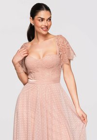 Femme aux cheveux foncés vêtue d'une robe en dentelle rose avec des manches bouffantes et une jupe en tulle à pois, souriante et tenant délicatement la jupe.
