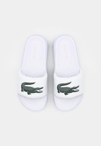 Sandale albe, cu talpă interioară texturată, având un logo verde de crocodil pe curea și un accent ușor violet pe talpa exterioară.