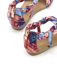 Sandále s kvetinovým vzorom, ktoré majú pletené popruhy, zips na päte, hnedú stielku a plochú podrážku. Farby zahŕňajú ružovú, modrú a fialovú.