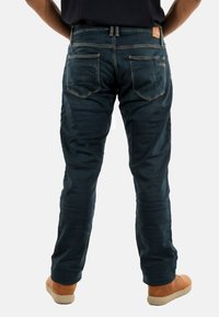Man in een donkerblauwe jeans en tan schoenen staat tegen een effen witte achtergrond, van achter gezien.