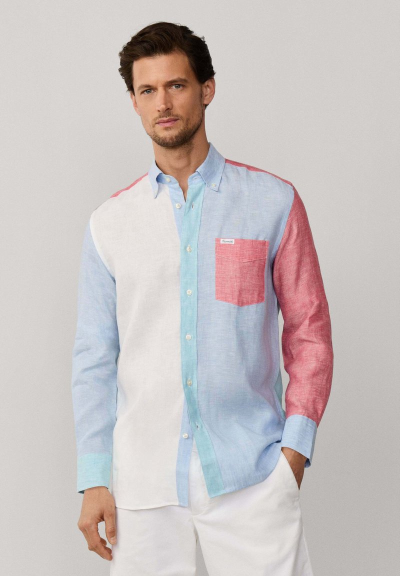Uomo che indossa una camicia a maniche lunghe con bottoni, caratterizzata da blocchi di colore bianco, azzurro chiaro, acqua e rosa, abbinata a pantaloni bianchi.