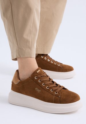 Zapatillas de ante marrón con suelas blancas gruesas y cordones marrones, combinadas con pantalones beige recortados sobre un fondo blanco.