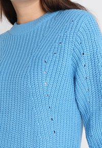 Vaaleansininen neulepaita, jossa on teksturoitu kuvio, pienillä silmäreikäkohdilla ja pyöreä kaula-aukko, valmistettu pehmeästä, paksusta langasta.