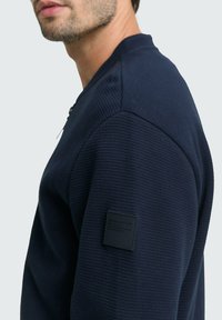 Felpa blu navy in tessuto testurizzato, con scollo rotondo e un patch quadrato nero sulla parte superiore della manica. Presenta una vestibilità atletica e dettagli cuciti.