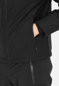 Sorte softshell-jakke med lynlåslommer og elastiske manchetter. Har et strømlinet design og glat tekstur, ideel til udendørsaktiviteter.