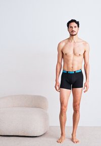 Fekete boxer alsónadrág kék derékkal, rajta a "Puma" logóval; a modell mezítláb áll egy minimalista környezetben.