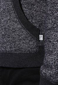 Sudadera de punto en negro y gris con cuello acanalado, tejido texturizado y una pequeña etiqueta de marca con un ícono en el lateral inferior.