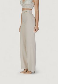 Pantaloni beige a vita alta, con gamba ampia e texture liscia, caratterizzati da due bottoni dorati frontali e una vestibilità sartoriale.