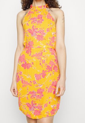 Robe jaune sans manches avec motif floral rose, col halter et taille cintrée, portée par une personne aux cheveux bouclés et avec des tatouages visibles sur les bras.