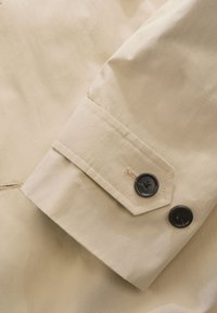 Manga de chaqueta beige con una textura suave. Presenta dos botones negros y un puño puntiagudo con costuras en contraste.