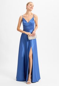 Blauwe satijnen jurk met spaghettibandjes, geraffineerd lijfje en een zijsplit. Model houdt een zilveren clutch vast en draagt zilveren sandalen met bandjes.