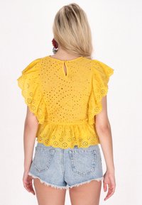Blusa gialla in pizzo con maniche corte a balze, vita a corpetto e dettaglio a apertura sulla schiena, abbinata a pantaloni shorts in denim sfrangiati.
