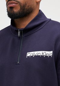 Granatowy polar zapinany na zamek z wysokim kołnierzem, z białym logo Calvin Klein na lewej piersi i teksturowanym materiałem.