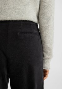 Zwarte broek met een gladde textuur, voorzien van een achterzak en subtiele stikdetails, gecombineerd met een lichtgrijze gebreide trui.