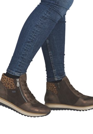 Persoon draagt donkerbruine enkellaarsjes met luipaardprint accenten en blauwe skinny jeans, staand met gekruiste benen.