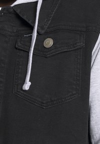 Czarna kamizelka z denimu z kieszenią na zatrzask na piersi, szarym ściągaczem i warstwową szarą tkaniną pod spodem. Tkanina o wyraźnej fakturze z widocznym przeszyciem.