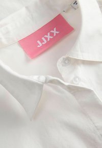 Bijela košulja s ovratnikom s ružičastom tkanom etiketom s brendom "JJXX" ušivenom unutrašnjosti vrata.