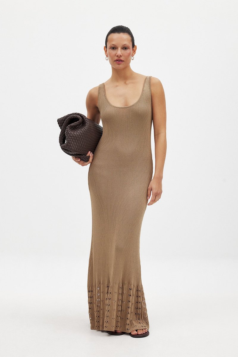 Maxi sukienka bez rękawów w kolorze beżowym, o prążkowanej strukturze i koronkowym wykończeniu. Modelka trzyma brązową, plecioną torebkę. Proste sandały dopełniają całość.