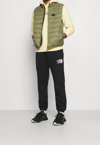 Olivgrön puffig väst över en ljusgul sweatshirt, parat med svarta sweatpants med en logotyp och svarta träningsskor.