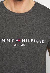 Primer plano de una persona que lleva una camiseta gris oscuro con el texto blanco "Tommy Hilfiger", el logotipo de la marca y "EST. 1985" bordado en el pecho.