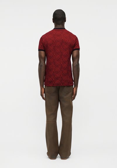 Hombre de pie de espaldas, vistiendo una camisa roja de manga corta con estampado y pantalones marrones de pierna recta, sobre un fondo liso.