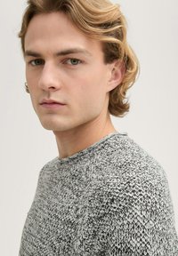 Gebreide trui in zwart en wit met een textured patroon; ronde hals en raglanmouwen; model poseert in een lichte hoek tegen een neutrale achtergrond.