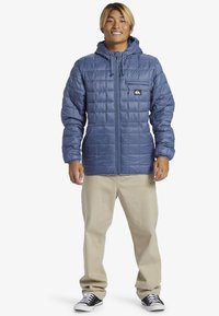 Gep quiltete blaue jacke mit kapuze, frontreißverschluss, brusttasche und seitentaschen. Kombiniert mit cremefarbenen hosen und schwarzen sneakers.