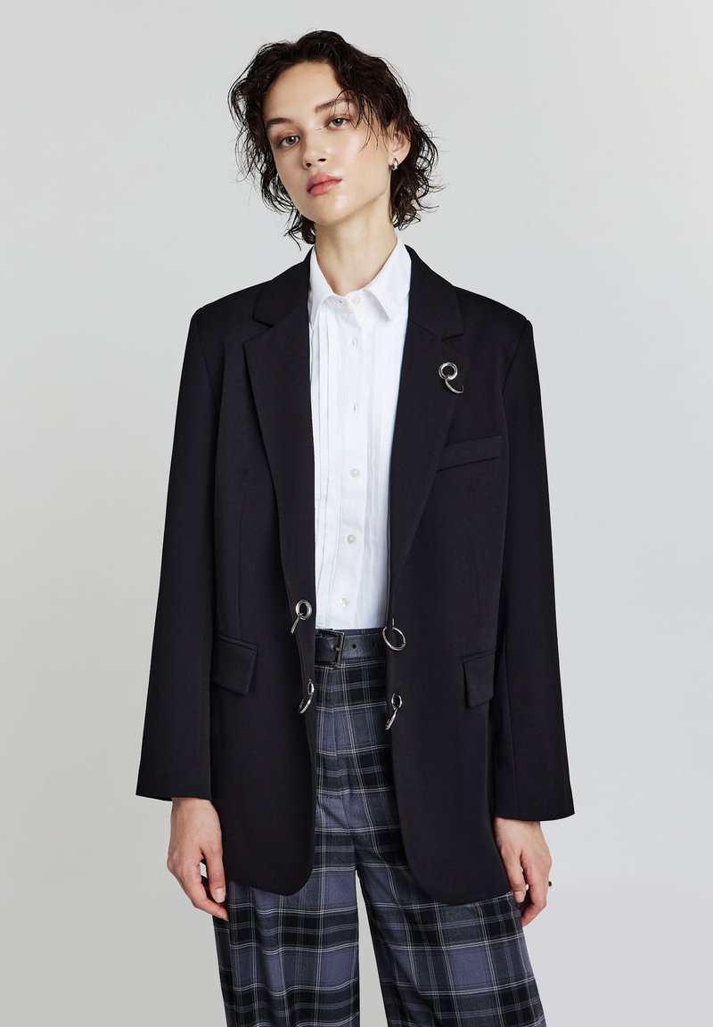 Blazer nero su misura con dettagli in metallo argentato, tasche frontali e revers a lancia, abbinato a una camicia bianca e pantaloni a quadri.