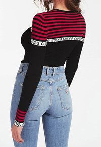 Mustat pitkähihaiset paidat, joissa on punaisia raitoja, ja merkkilogoisa joustovyö. Vaaleansiniset korkeavyötäröiset farkut, joissa on sileä pinta ja taskut.